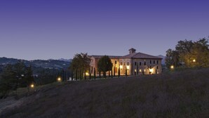 Un grand bâtiment en pierre, doté de nombreuses fenêtres et d’une tour centrale, entouré d’arbres et situé dans un paysage vallonné.