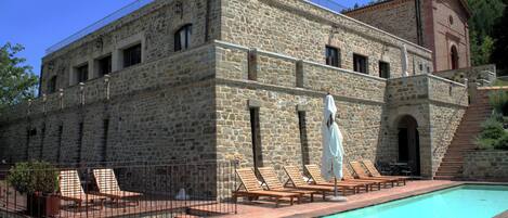 A stone building with a swimming pool, wooden lounge chairs, and a staircase leading to a higher level.