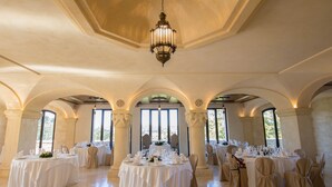 Une salle de banquet avec des tables rondes disposées pour un événement formel, dotée d’un plafond haut et de fenêtres en arc de cercle.