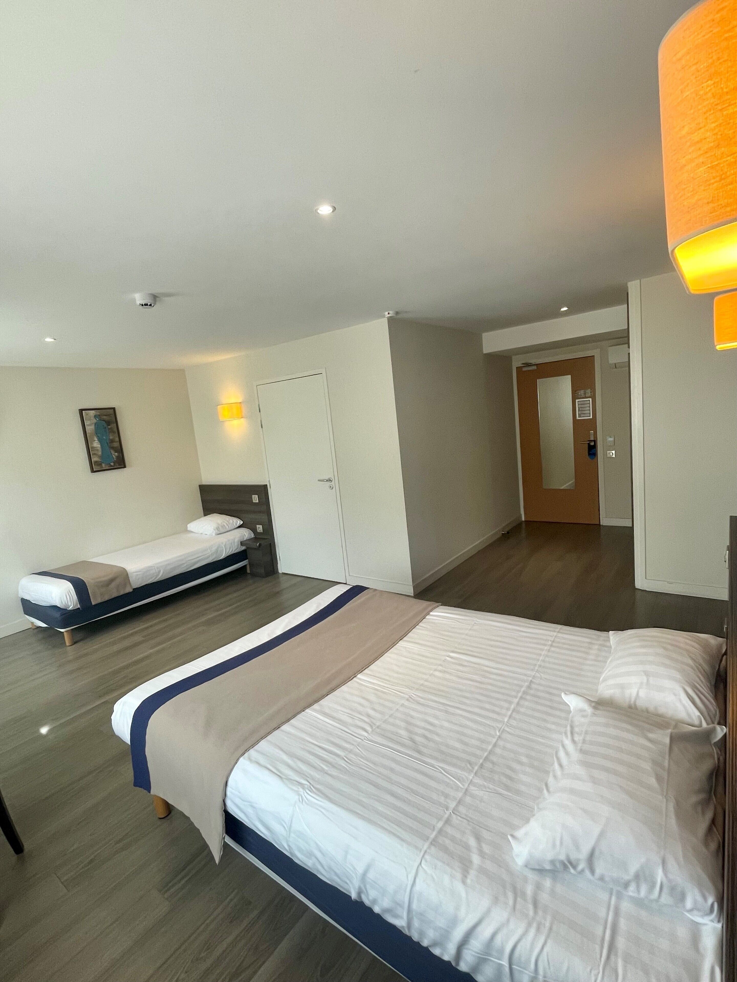 A hotel room with a large bed, a smaller bed, a wardrobe, a door, and a painting on the wall.