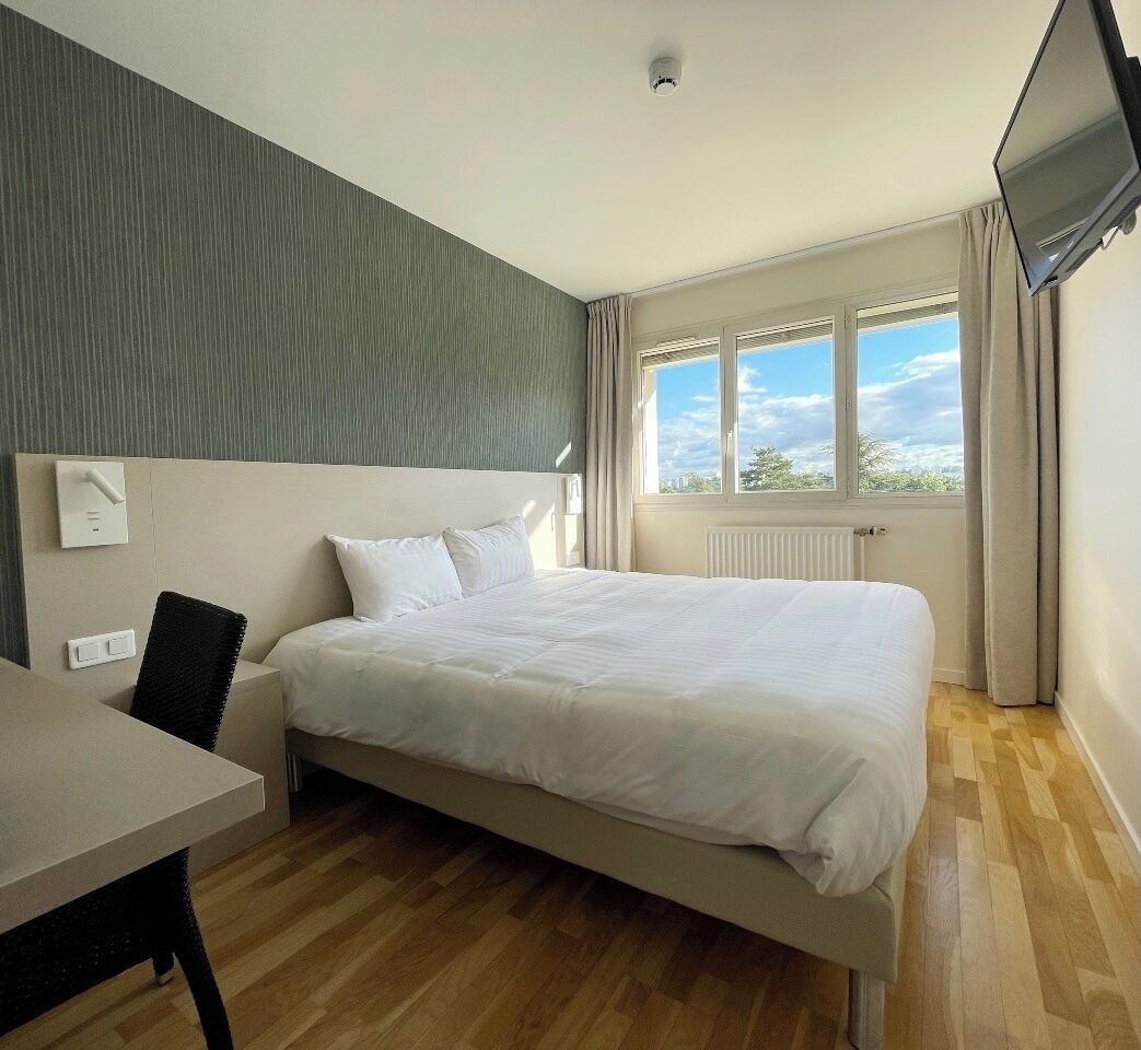 A hotel room with a bed, a desk, a chair, a window with curtains, and a flat-screen TV mounted on the wall.