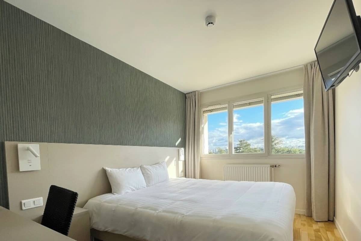 A hotel room with a bed, a desk, a chair, a window with curtains, and a flat-screen TV mounted on the wall.