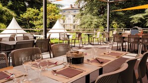 An outdoor dining area with tables and chairs, a large umbrella, and a building in the background. - Valpré Lyon Hôtel (Ecully)