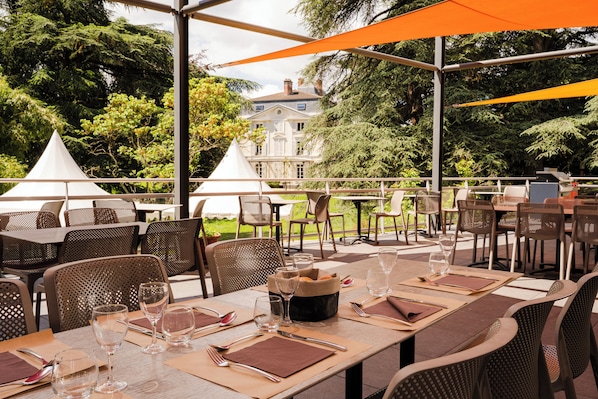 An outdoor dining area with tables and chairs, a large umbrella, and a building in the background. - Valpré Lyon Hôtel (Ecully)