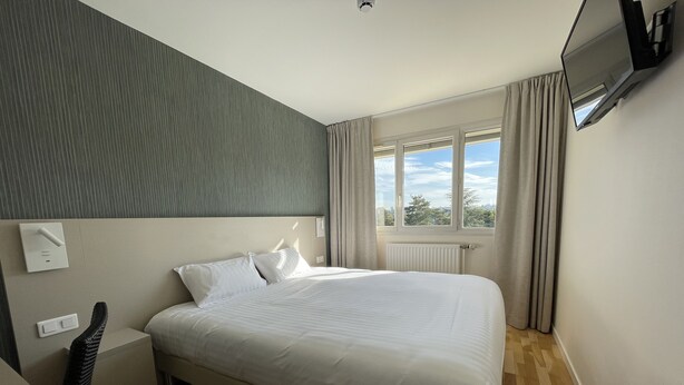 Une chambre d  ’hôtel moderne avec un grand lit, une table de chevet, une chaise, un téléviseur à écran plat fixé au mur et une fenêtre avec des rideaux.