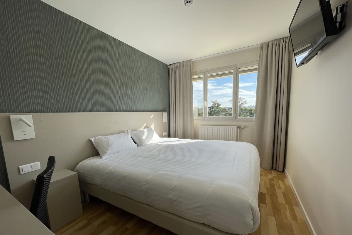 A modern hotel room with a large bed, a bedside table, a chair, a flat-screen TV mounted on the wall, and a window with curtains.