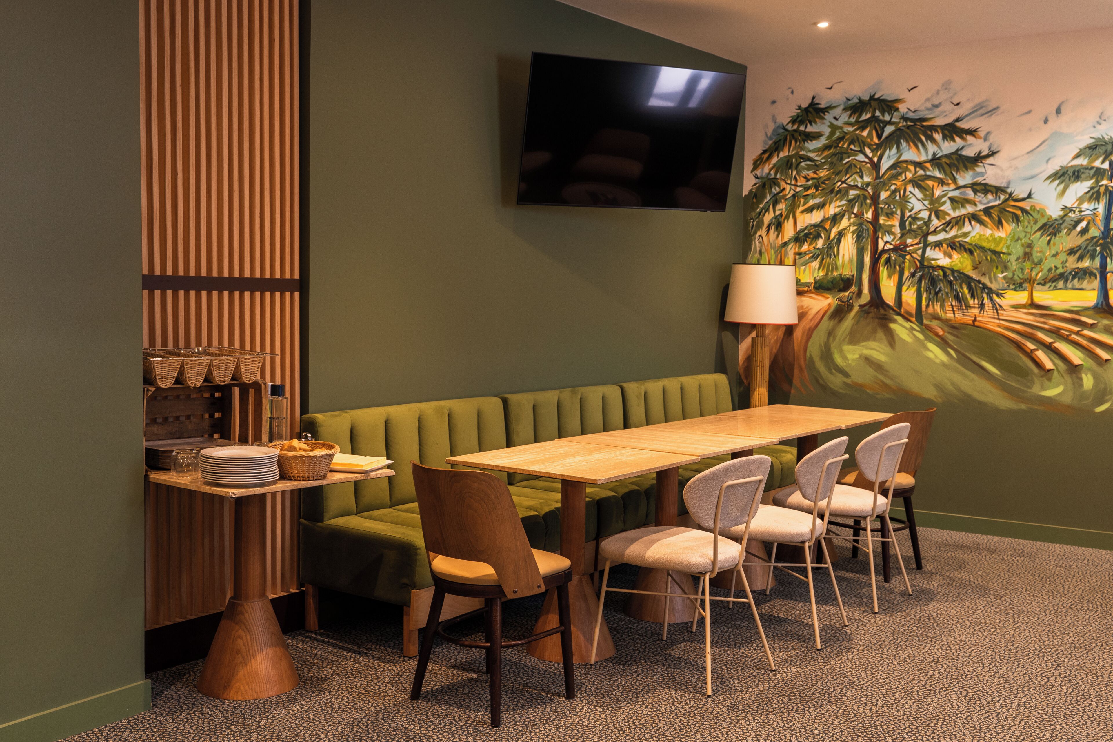 A dining area with a green booth, wooden table, and chairs, a flat-screen TV mounted on the wall, and a painting of a tropical scene.