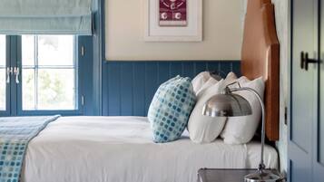 Une chambre à coucher avec un lit, une fenêtre avec un store bleu, une tête de lit, un tableau accroché au mur et une petite commode en bois.