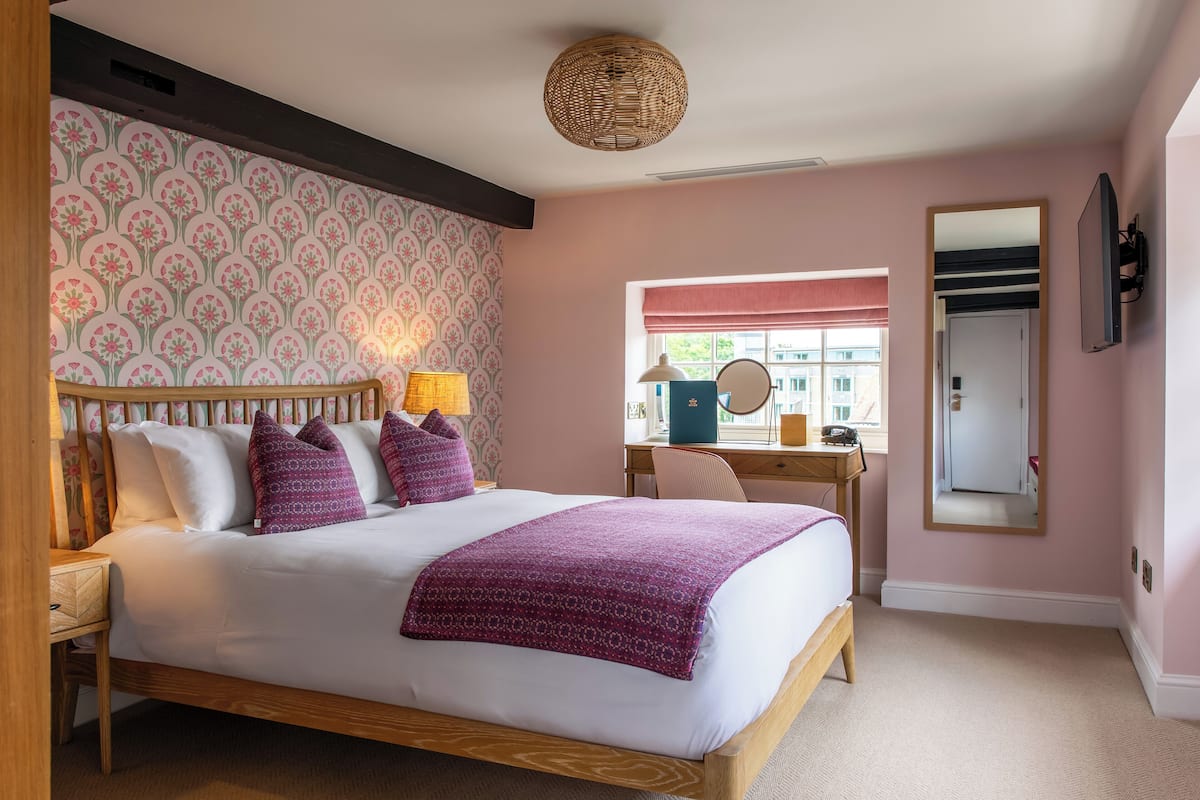 A bedroom with a bed, bedside table, lamp, dresser, mirror, and window with a view.