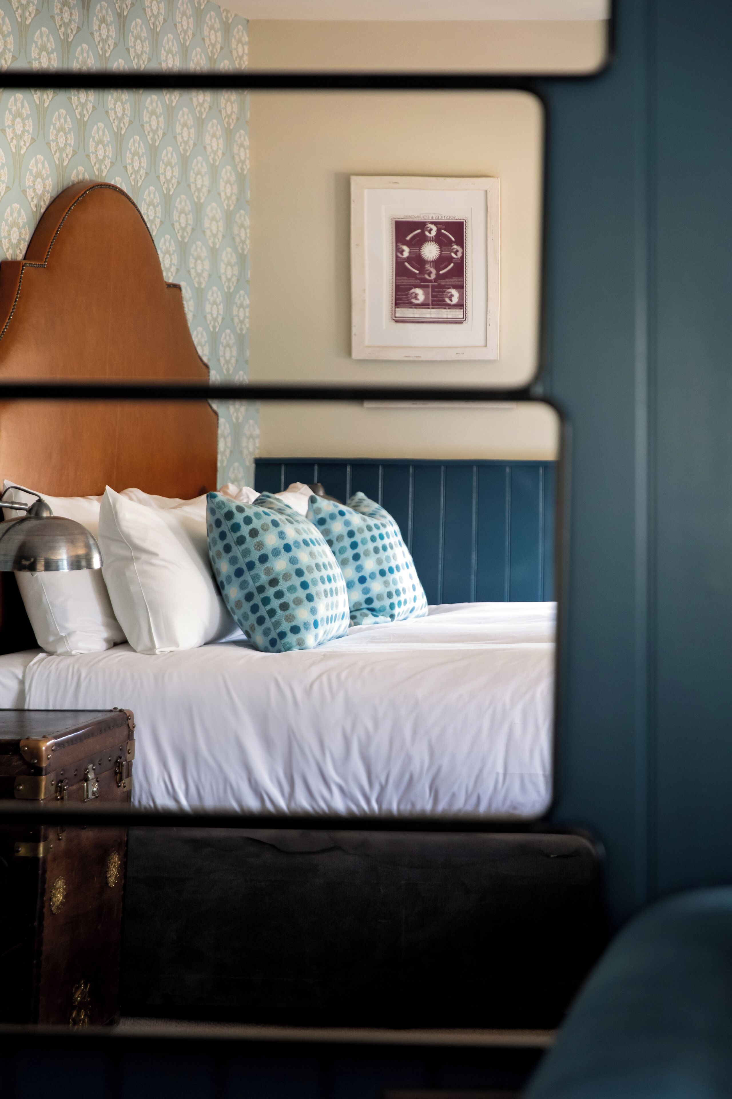 Una cama con cabecera de madera, ropa de cama blanca y almohadas estampadas. Un maletero antiguo se ve en el suelo.