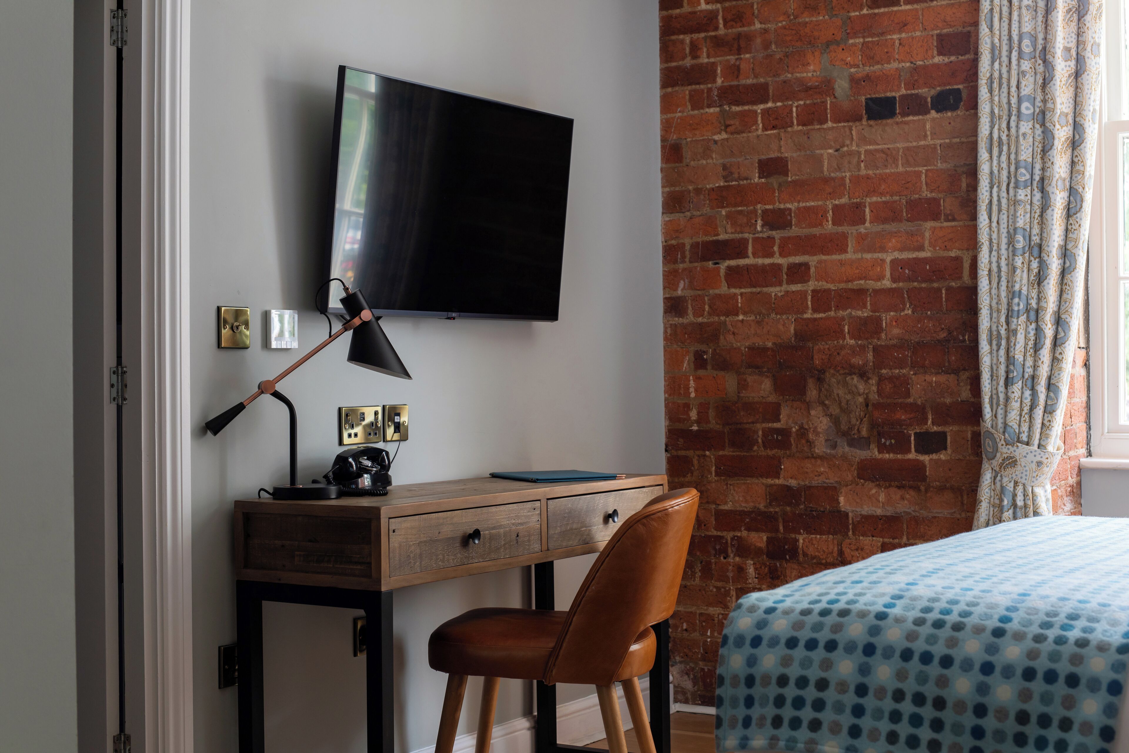 A room with a brick wall, a flat-screen TV mounted above a wooden desk, a brown leather chair, and a bed with a blue polka dot bedspread.