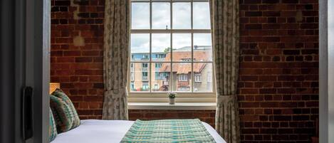 A room with a brick wall, a large window with curtains, a bed with a patterned blanket, and a small plant on the windowsill.
