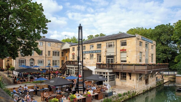 Ein Restaurant am Flussufer mit Terrassenplätzen, einem Schild mit der Aufschrift "The Head of the River" und einer Brücke, die zu einem anderen Gebäude führt.