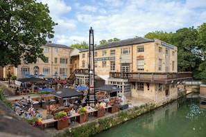 Un restaurante junto al río con mesas al aire libre, un cartel que dice "The Head of the River" y un puente que conecta con otro edificio.