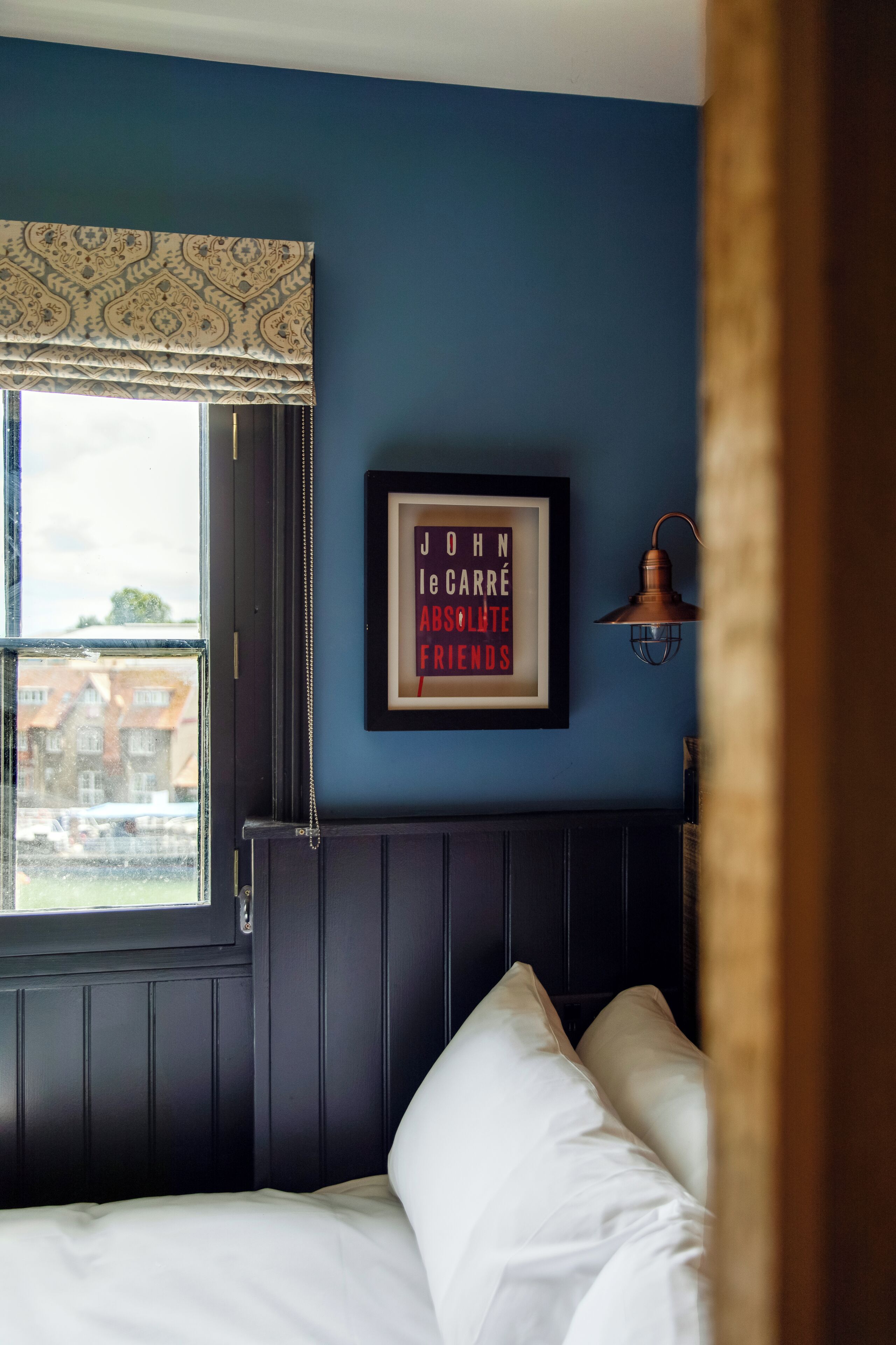 A room with a bed, a framed quote on the wall, a window with a patterned valance, and a wall-mounted lamp.