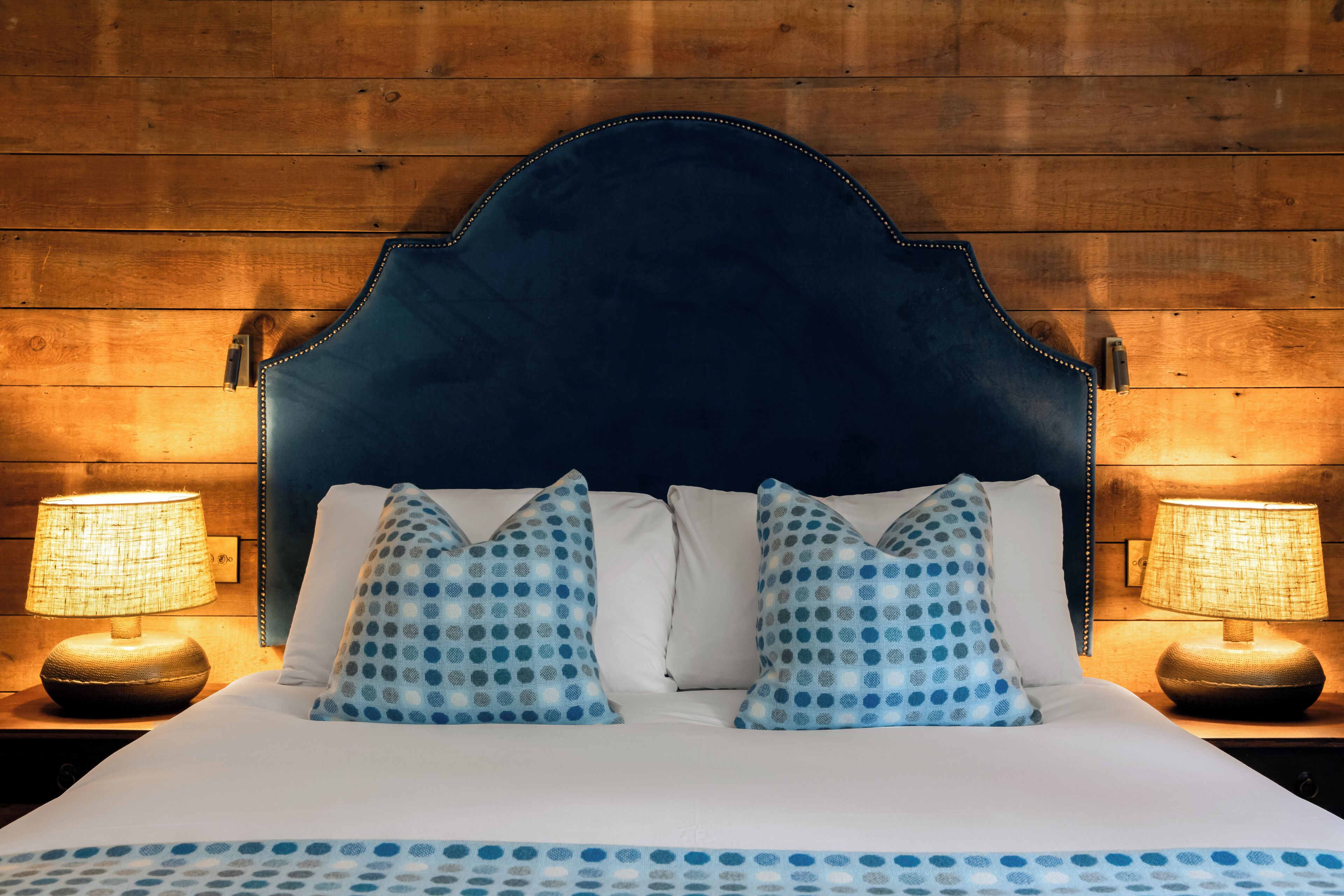 A bed with a dark headboard, white bedding, and blue polka dot pillows. There are two bedside lamps on wooden nightstands.