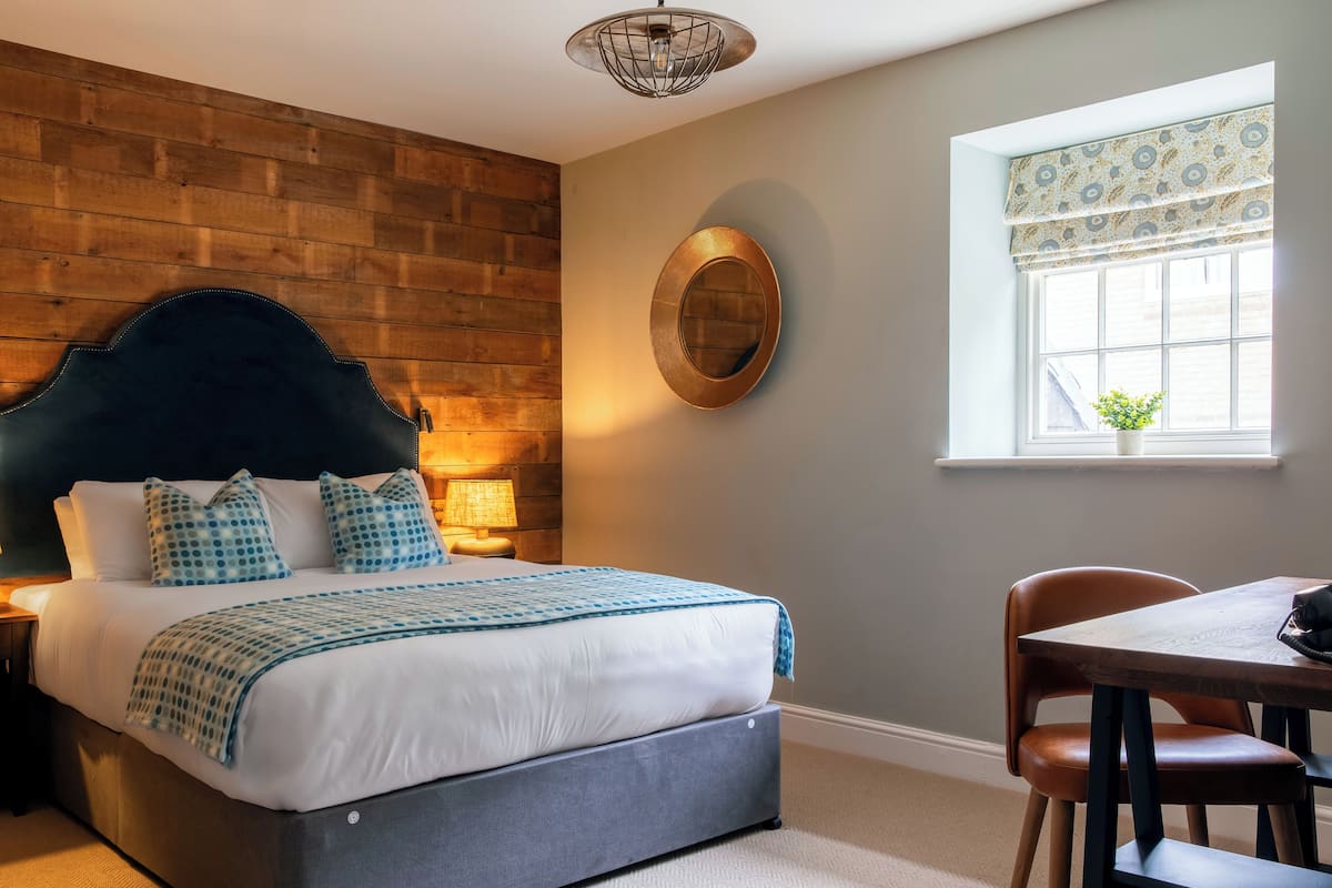 A bedroom with a bed, a wooden wall, a window with curtains, a table with a chair, and a hanging light fixture.