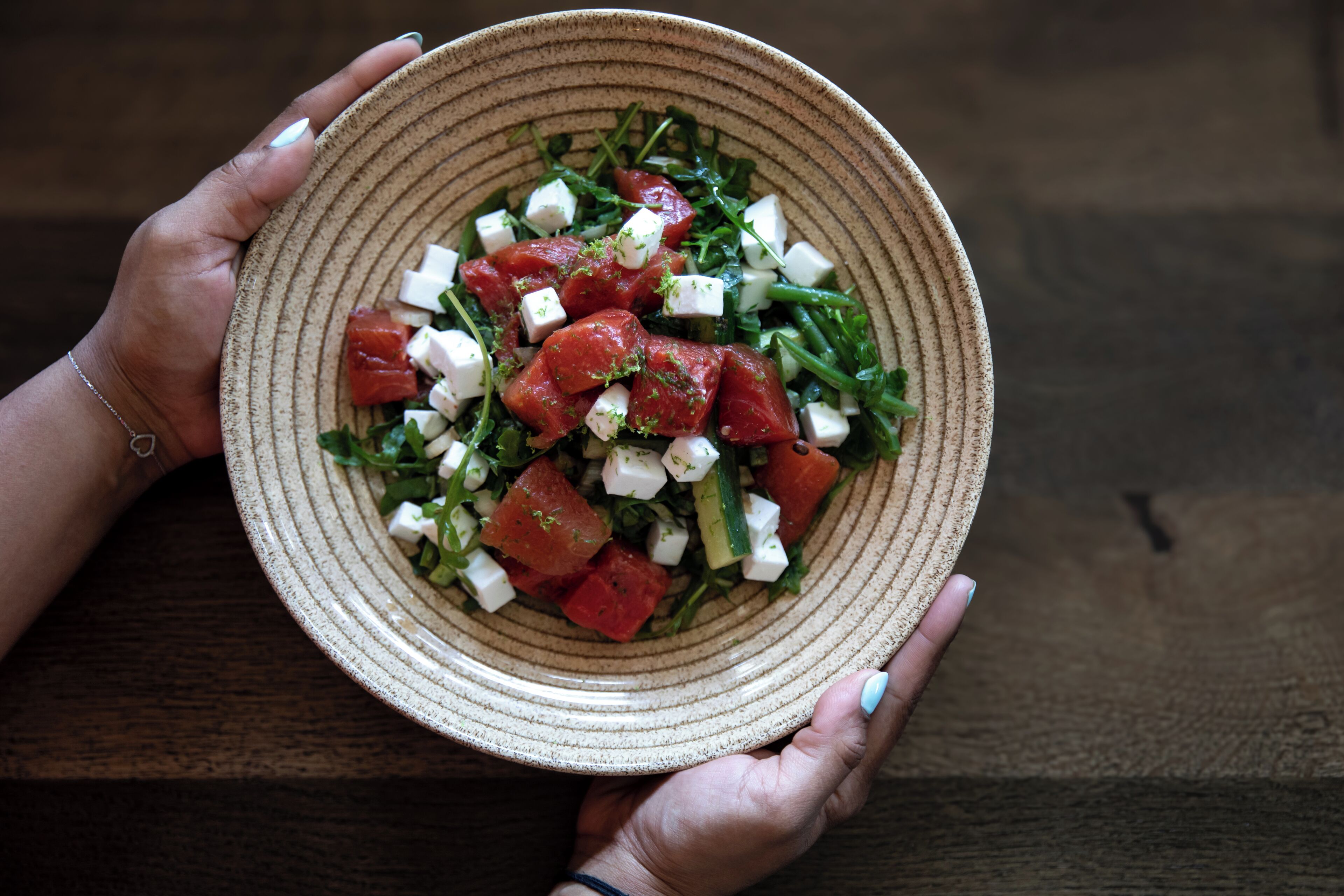 A person holding a bowl of salad with tomatoes, cucumbers, and cheese.