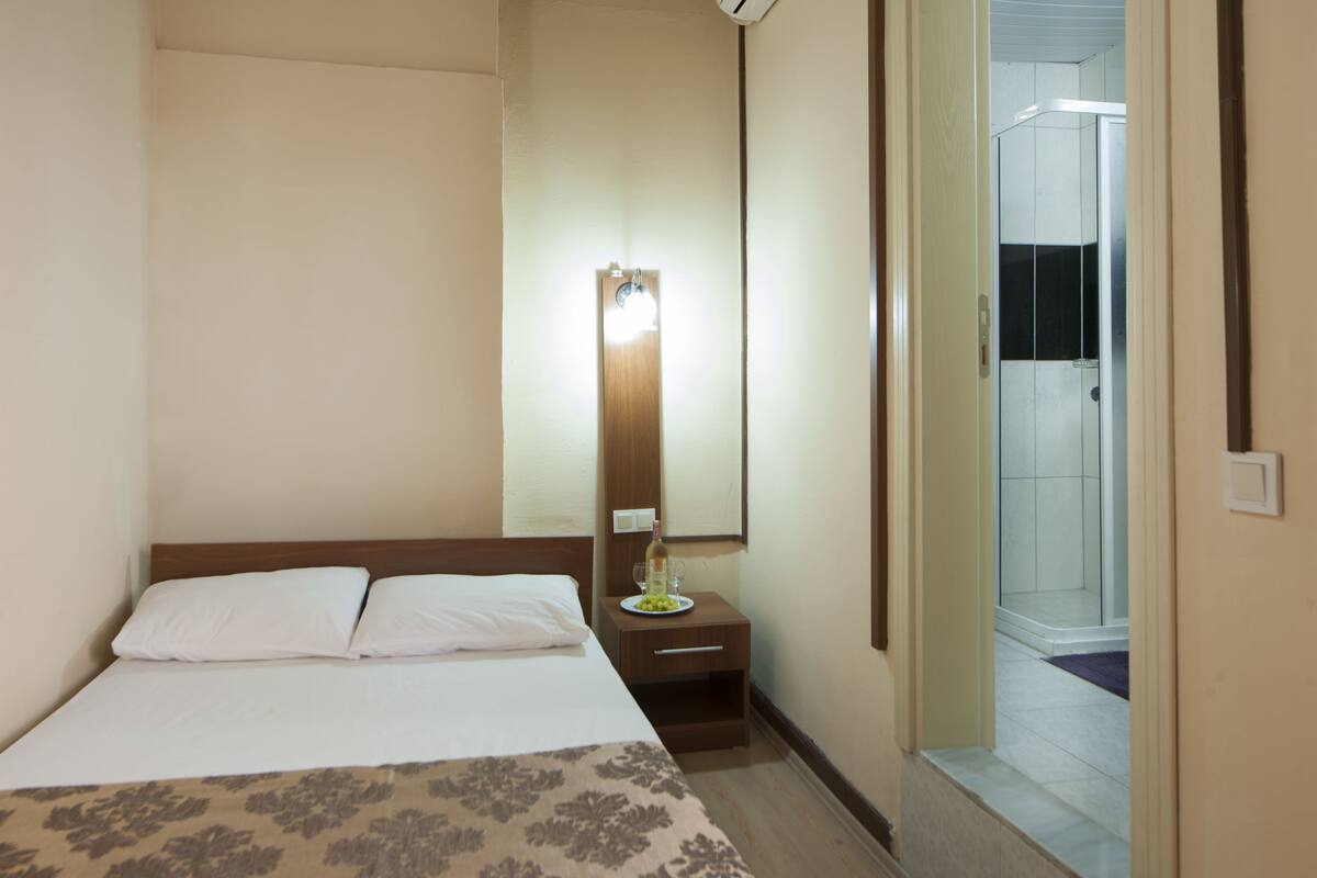 A hotel room with a bed, a nightstand with a bottle and glasses, a mirror, and a shower area visible through an open door.