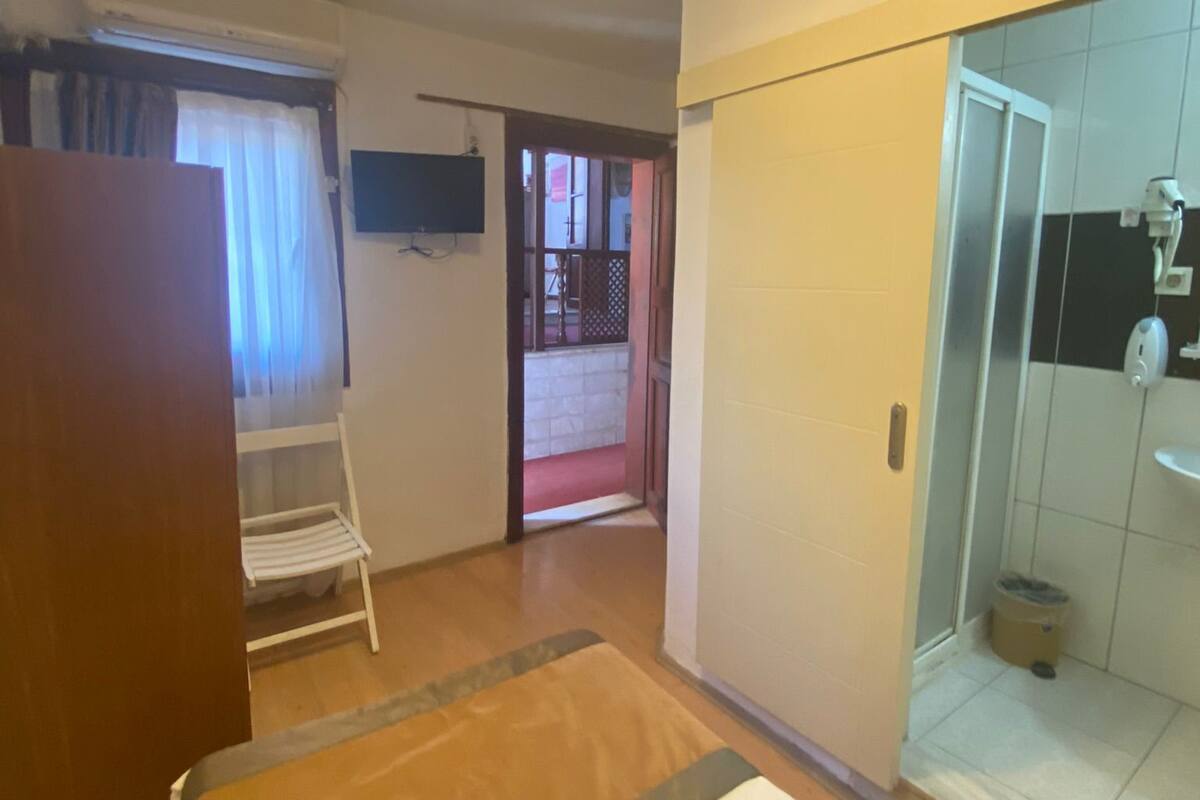 A hotel room with a bed, a wardrobe, a small table, a chair, a shower area, and a view of the outside.
