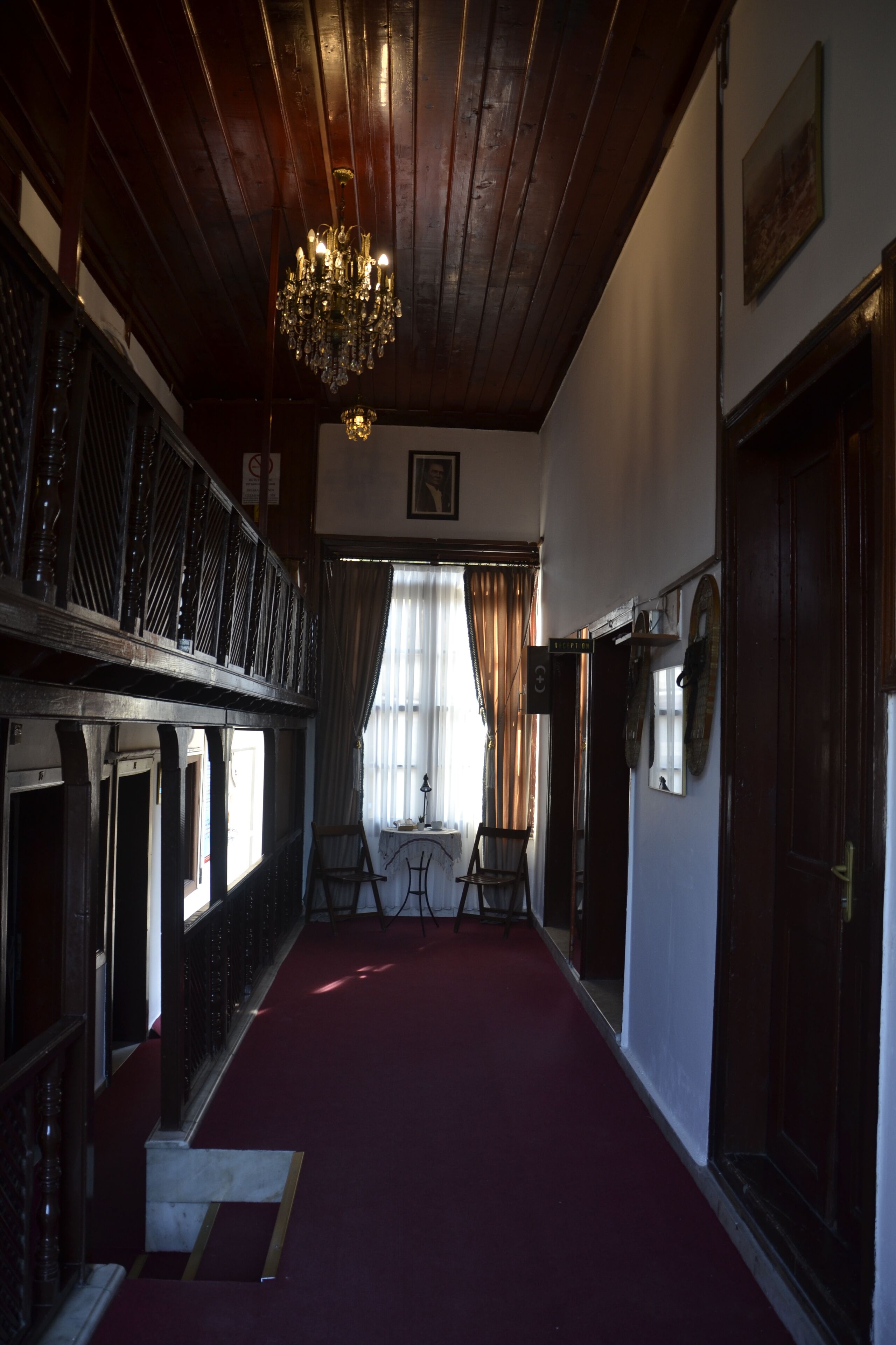 Een hal met een rood tapijt, houten plafond en een kroonluchter. Aan de rechterkant bevinden zich twee deuren en aan de linkerkant een raam met gordijnen.