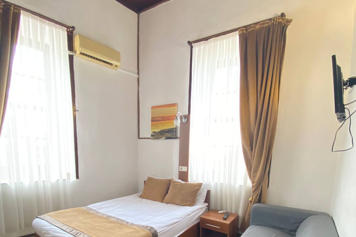 A room with a bed, a grey sofa, wooden flooring, a painting on the wall, and a window with curtains.
