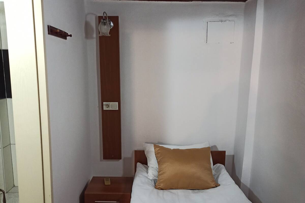 A small, neatly arranged bedroom with a wooden headboard, a single bed with white and brown bedding, a nightstand, and a wall-mounted light fixture.