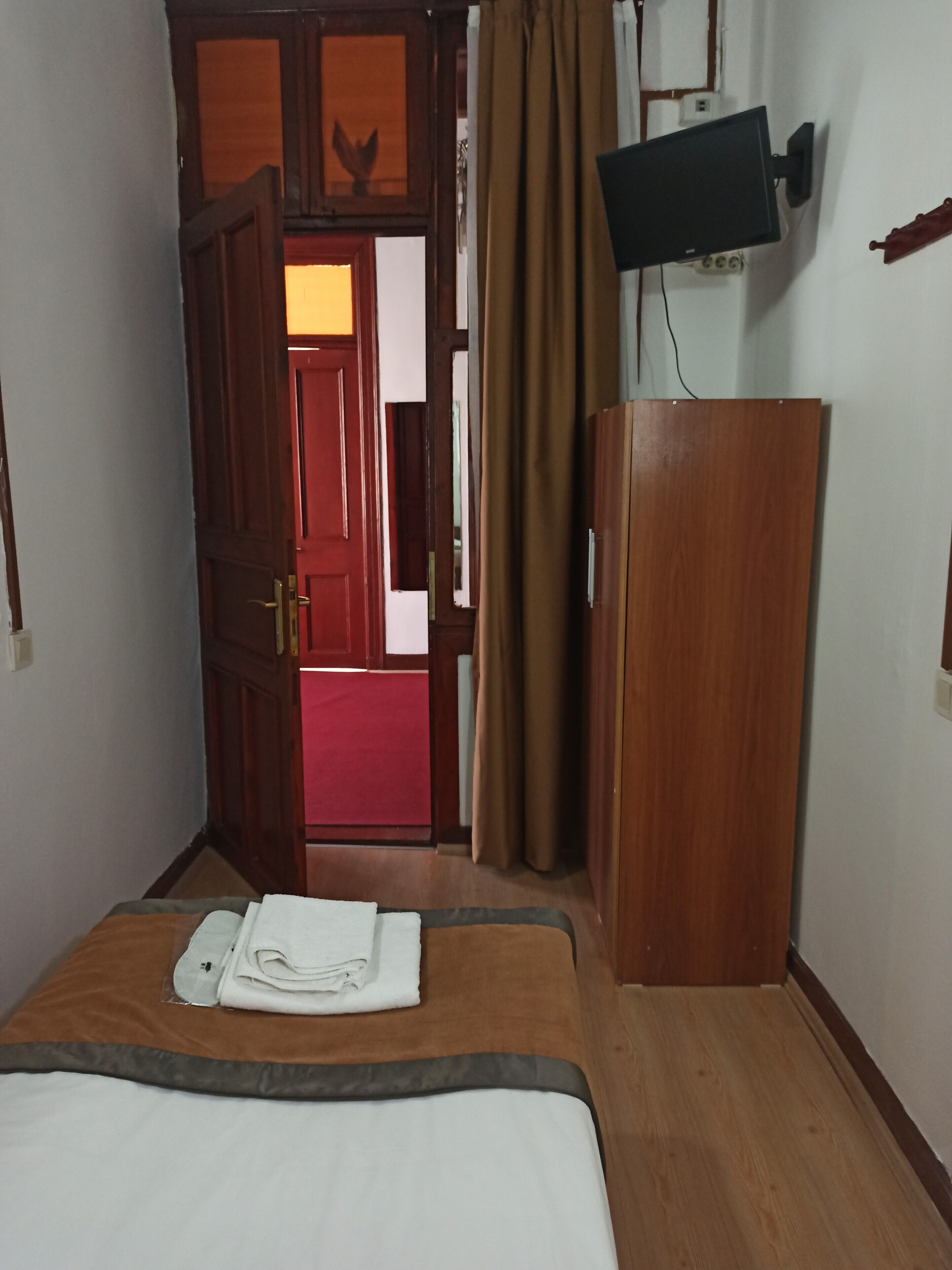 A hotel room with a bed, a wooden wardrobe, a television mounted on the wall, and a red door leading to another room.