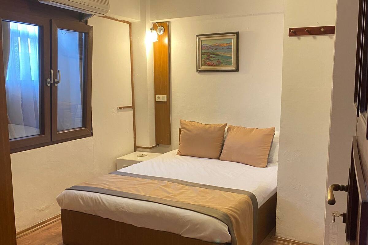 A bedroom with a bed, wooden flooring, a window with curtains, a wall-mounted air conditioner, and a painting.