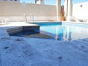 Una piscina con solárium de baldosas, un espacio pequeño junto a la piscina con sillas y una planta en maceta.
