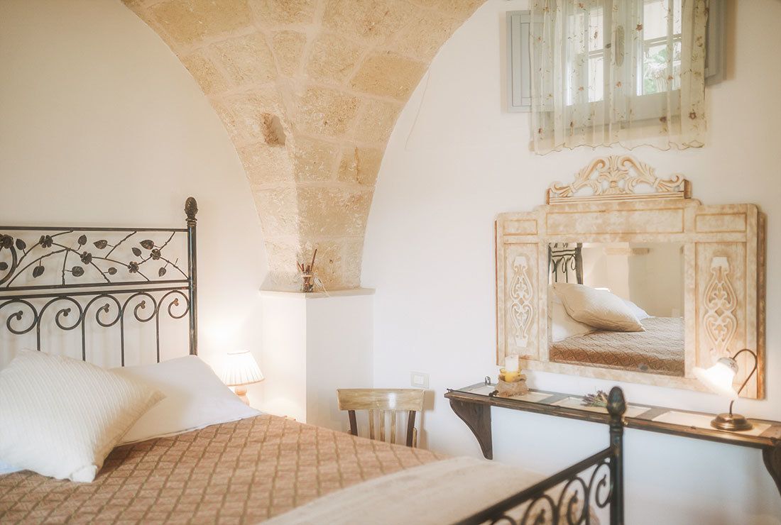 Une chambre avec une arche en pierre, un lit recouvert d’une couvre-lit à motifs, une table de chevet avec une lampe et un miroir doté d’un cadre décoratif.
