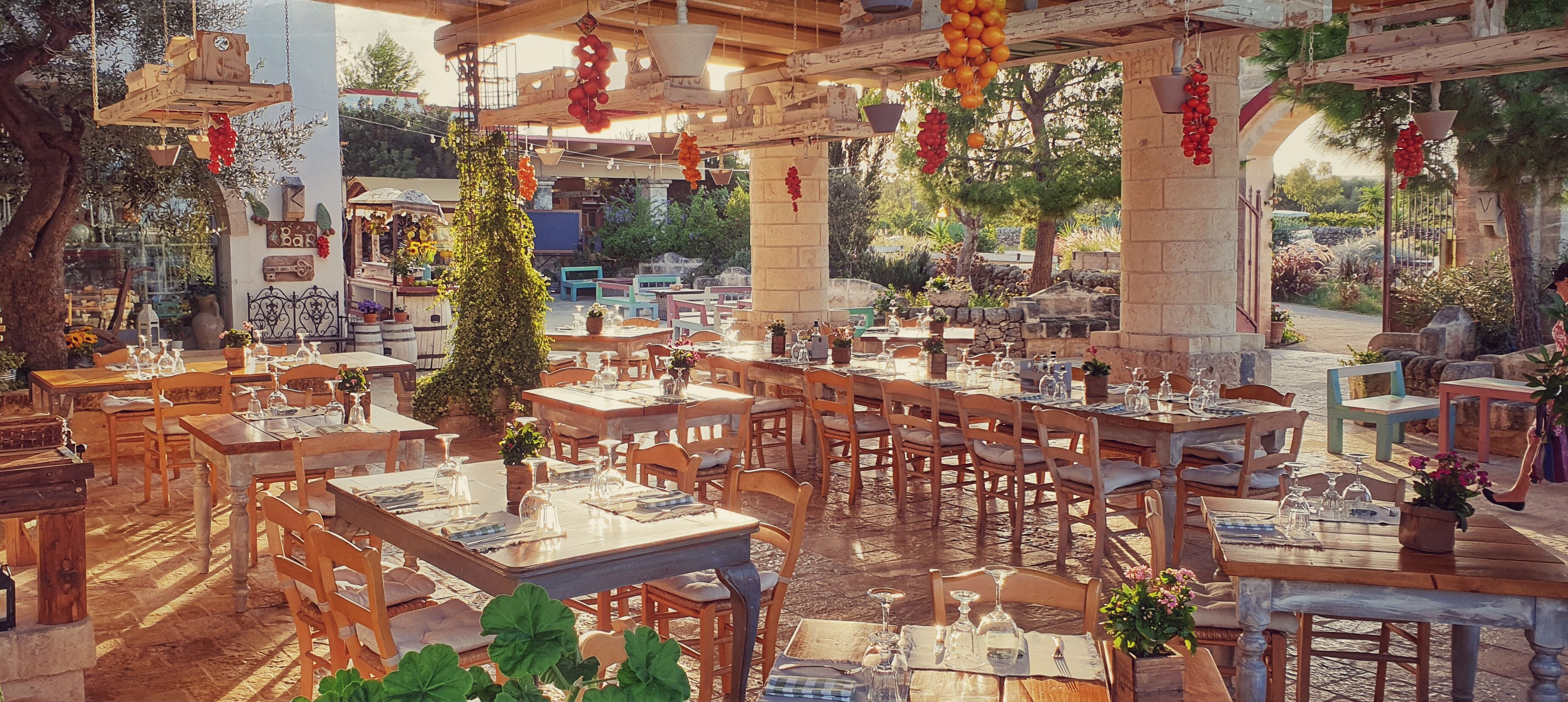 An outdoor restaurant with multiple wooden tables and chairs, surrounded by stone walls and greenery.