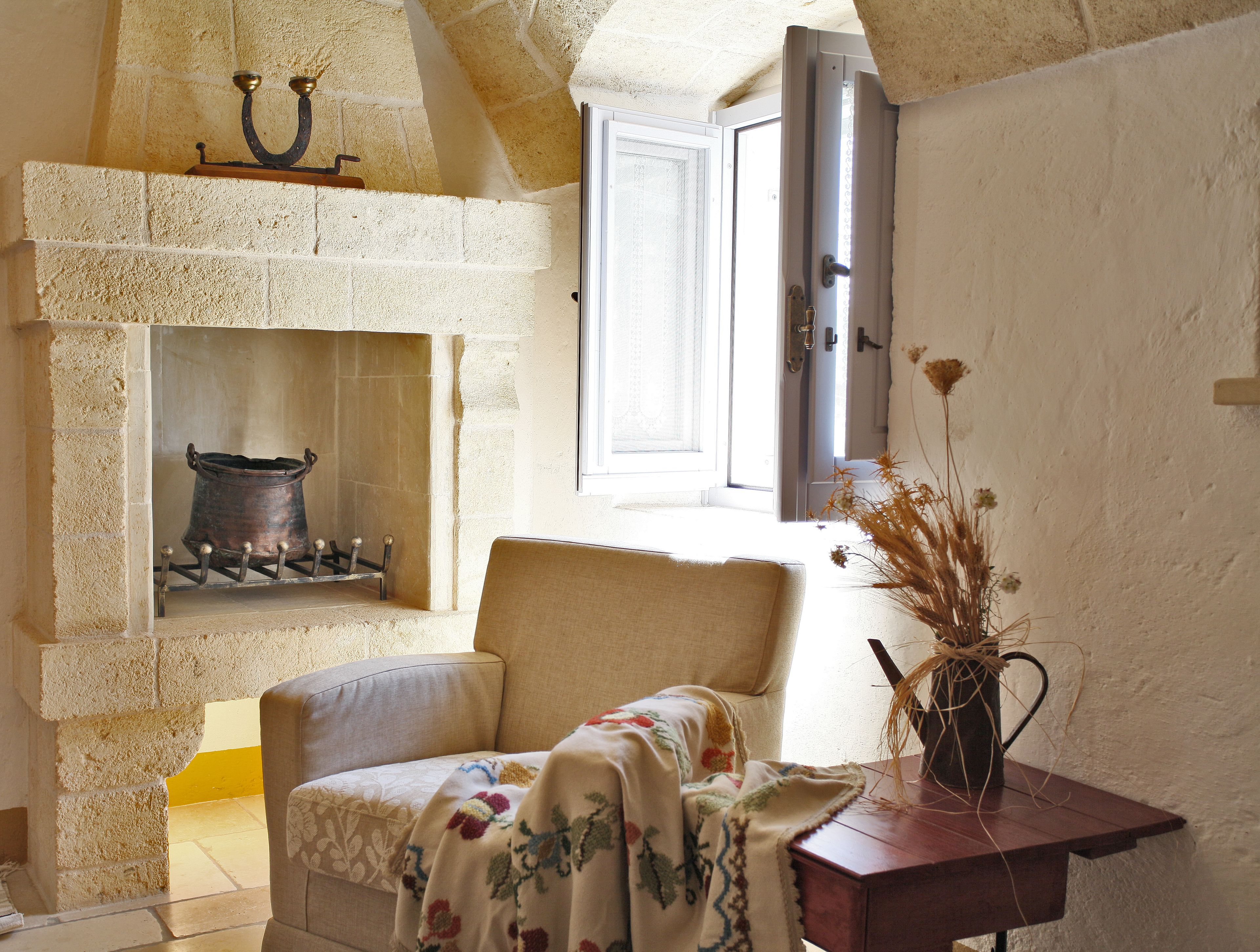 A cozy room with a stone fireplace, a comfortable chair, a wooden table with a vase, and a window with a view.