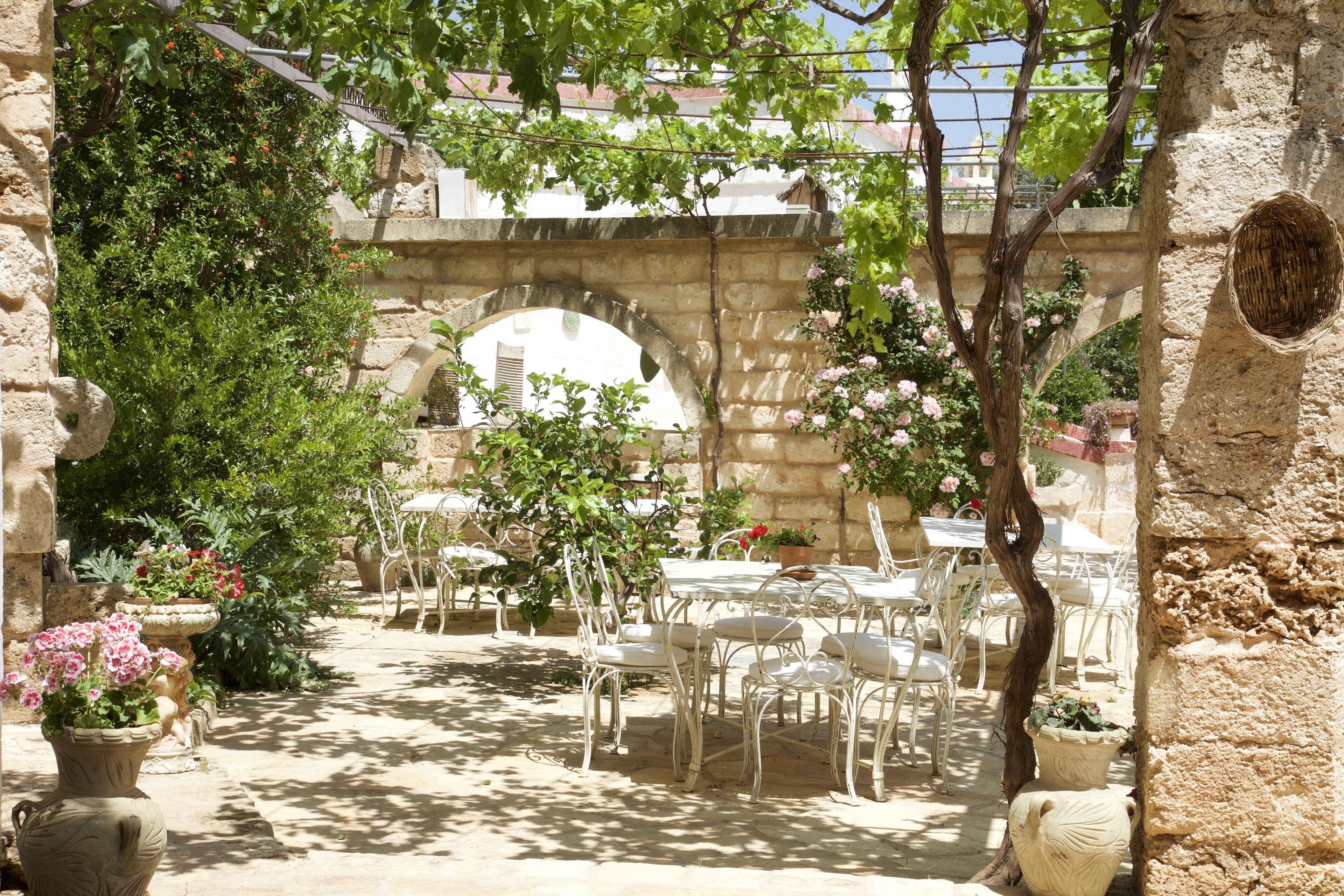 An outdoor patio with stone arches, white metal chairs, and potted plants.
