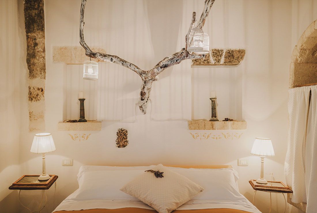 A bedroom with a large bed, two bedside tables with lamps, a decorative branch wall art piece, and a window with curtains.