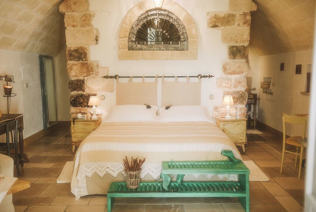 Un dormitorio acogedor con una pared de piedra, una cama con cabecera de madera, dos mesitas de noche con lámparas y un banco verde.