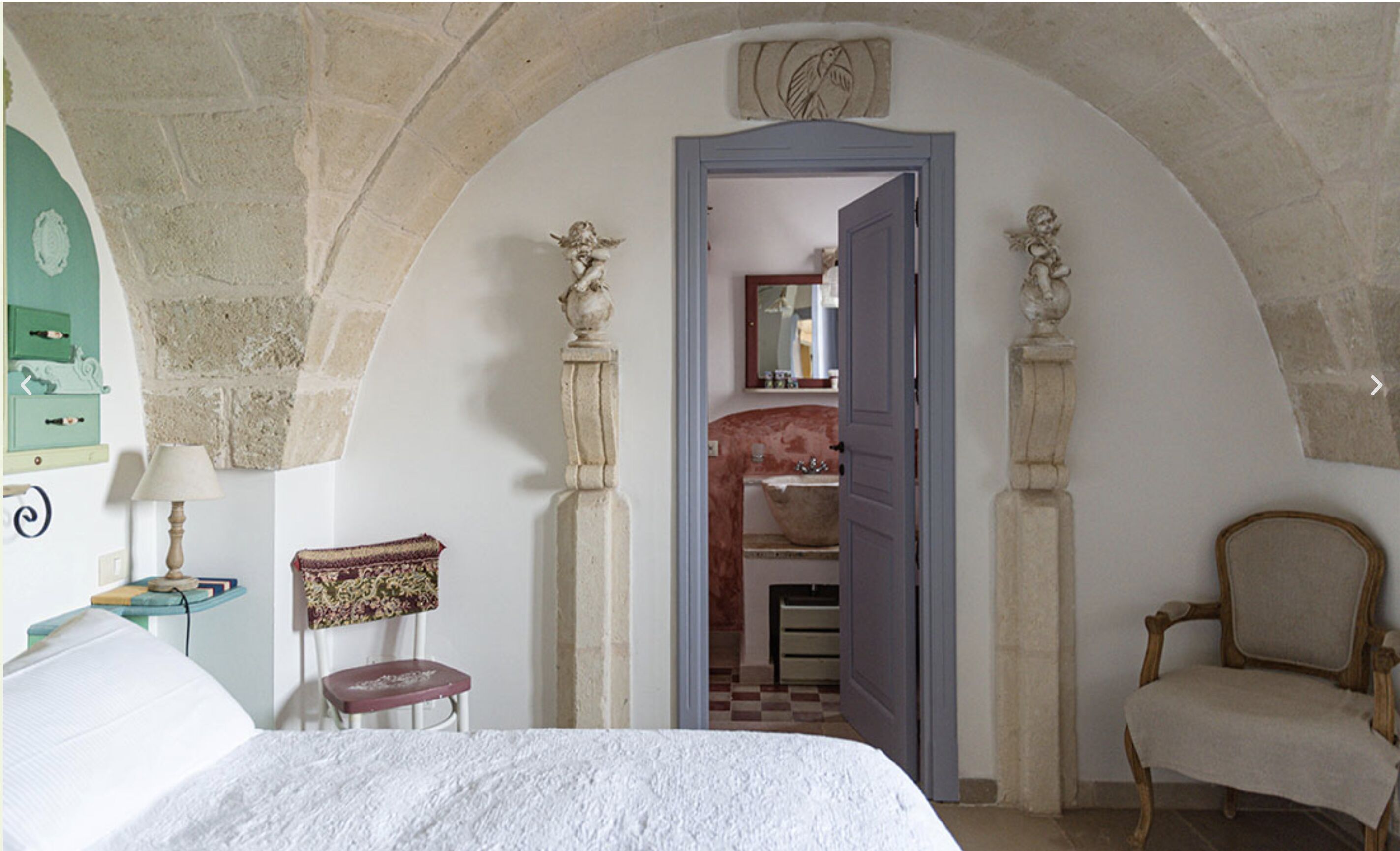 A bedroom with a bed, a chair, a small table with a lamp, and a mirror.