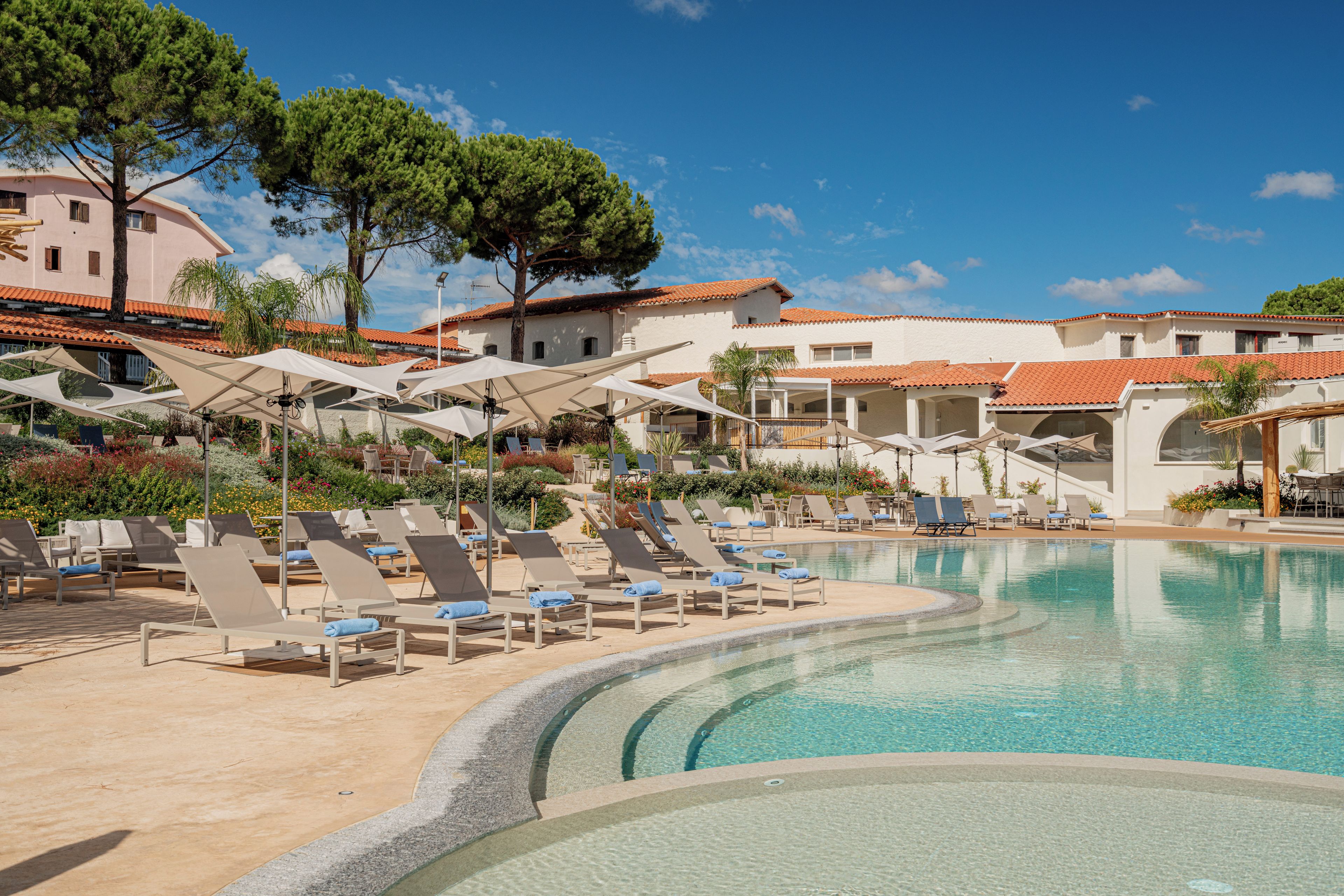 A poolside area with lounge chairs and umbrellas, surrounded by buildings with red-tiled roofs and a clear blue sky.