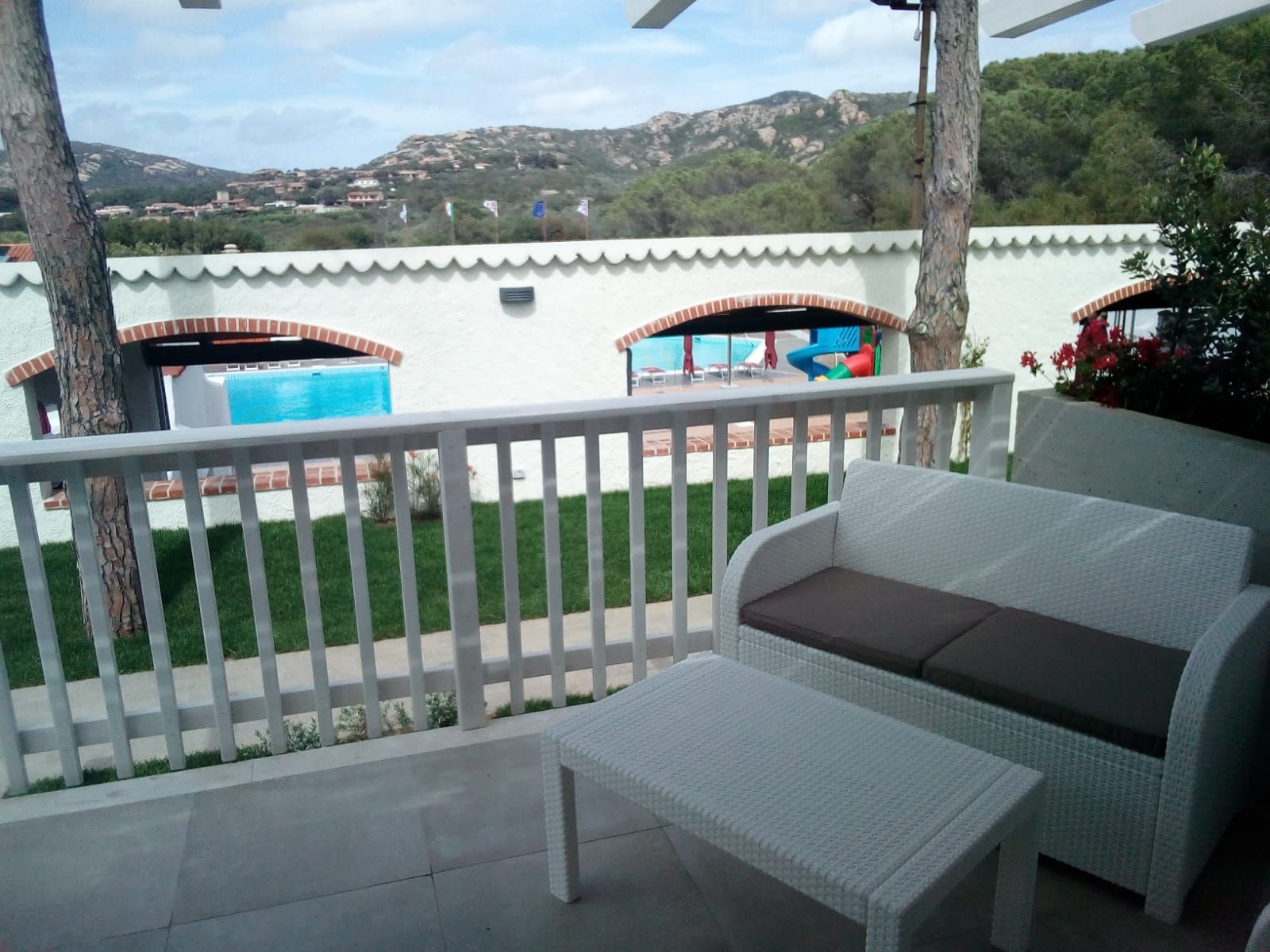 A covered outdoor area with a white wicker sofa, a small table, and a view of a swimming pool and hills in the background.
