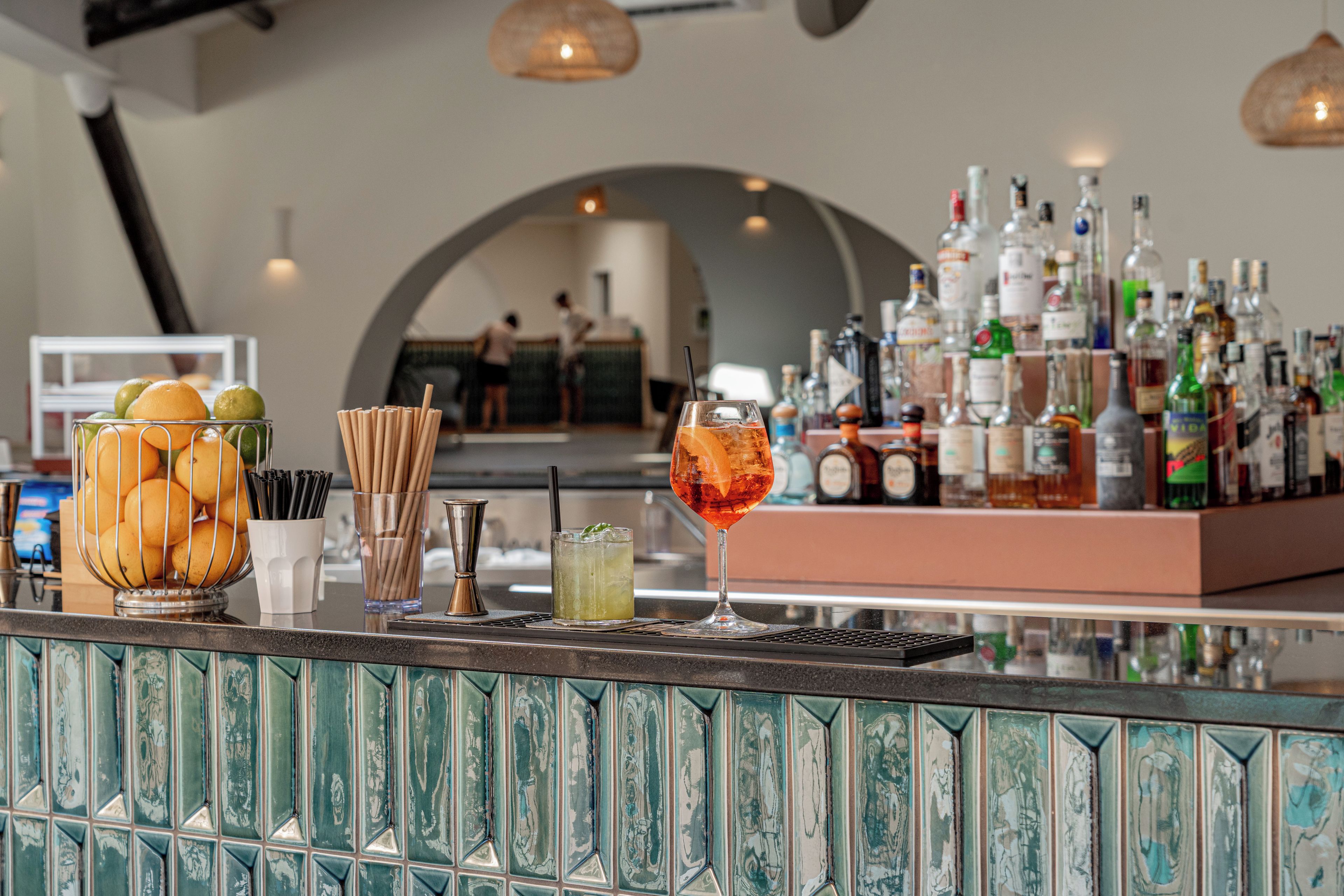 Un coin bar avec un assortiment de bouteilles et de verres, un panier de fruits et un mur décoratif orné d’un motif géométrique.