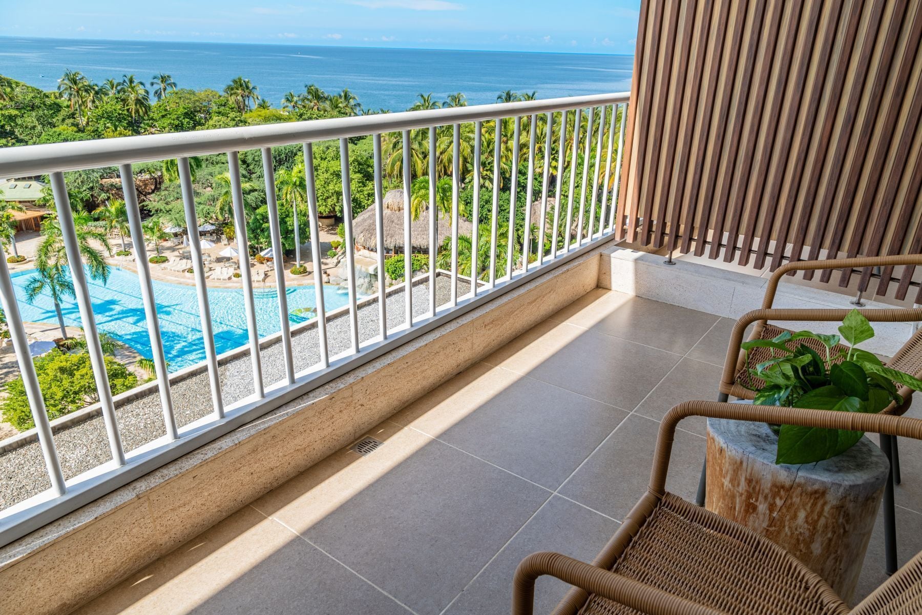 A balcony with a view of a pool and ocean, wicker furniture, and a stone column.
