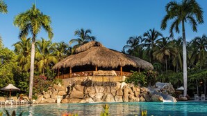 A resort pool with a thatched-roof structure, palm trees, and a rocky island. - Hotel Irotama Lago (Santa Marta)