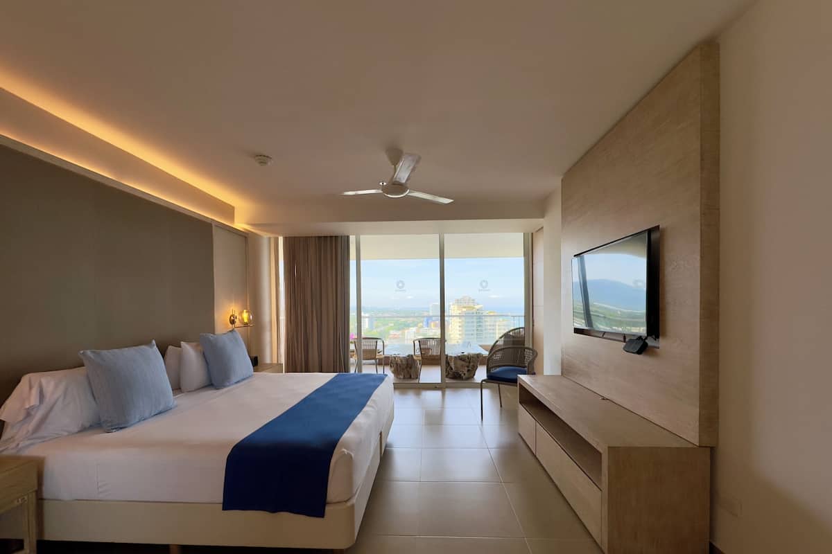 A modern hotel room with a large bed, a TV mounted on the wall, and a balcony with a view of the city.