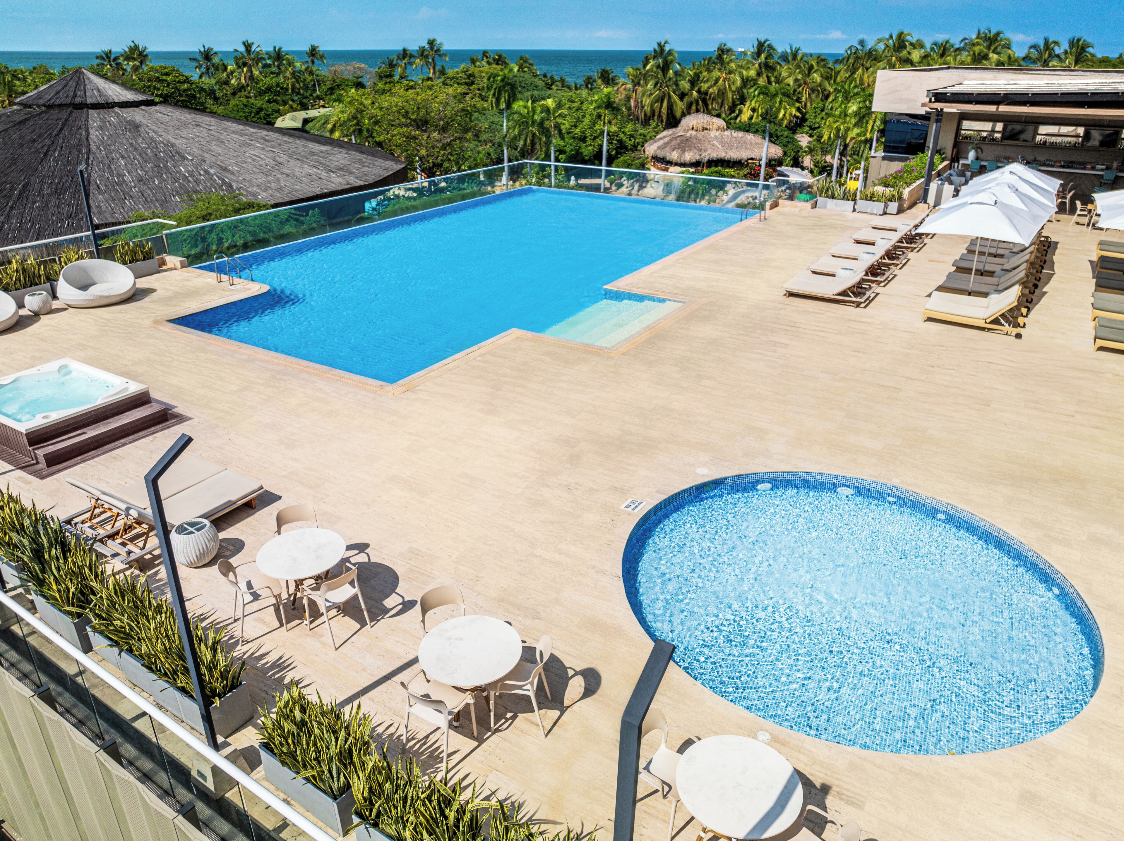 A large rectangular pool with a smaller circular pool, surrounded by lounge chairs and tables, with a view of the ocean and palm trees.