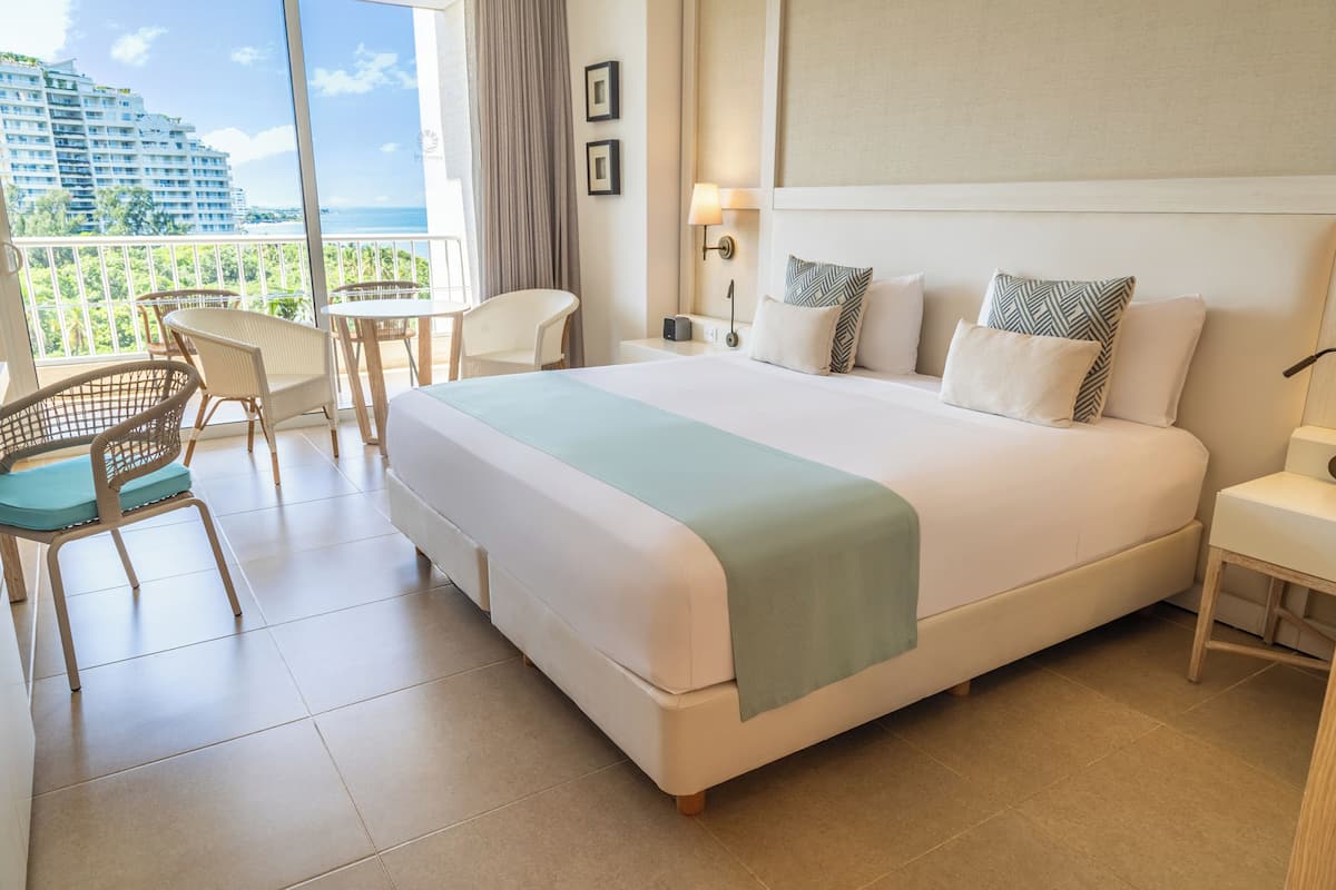 A hotel room with a large bed, a balcony with a view of the city, a small table, and a chair.