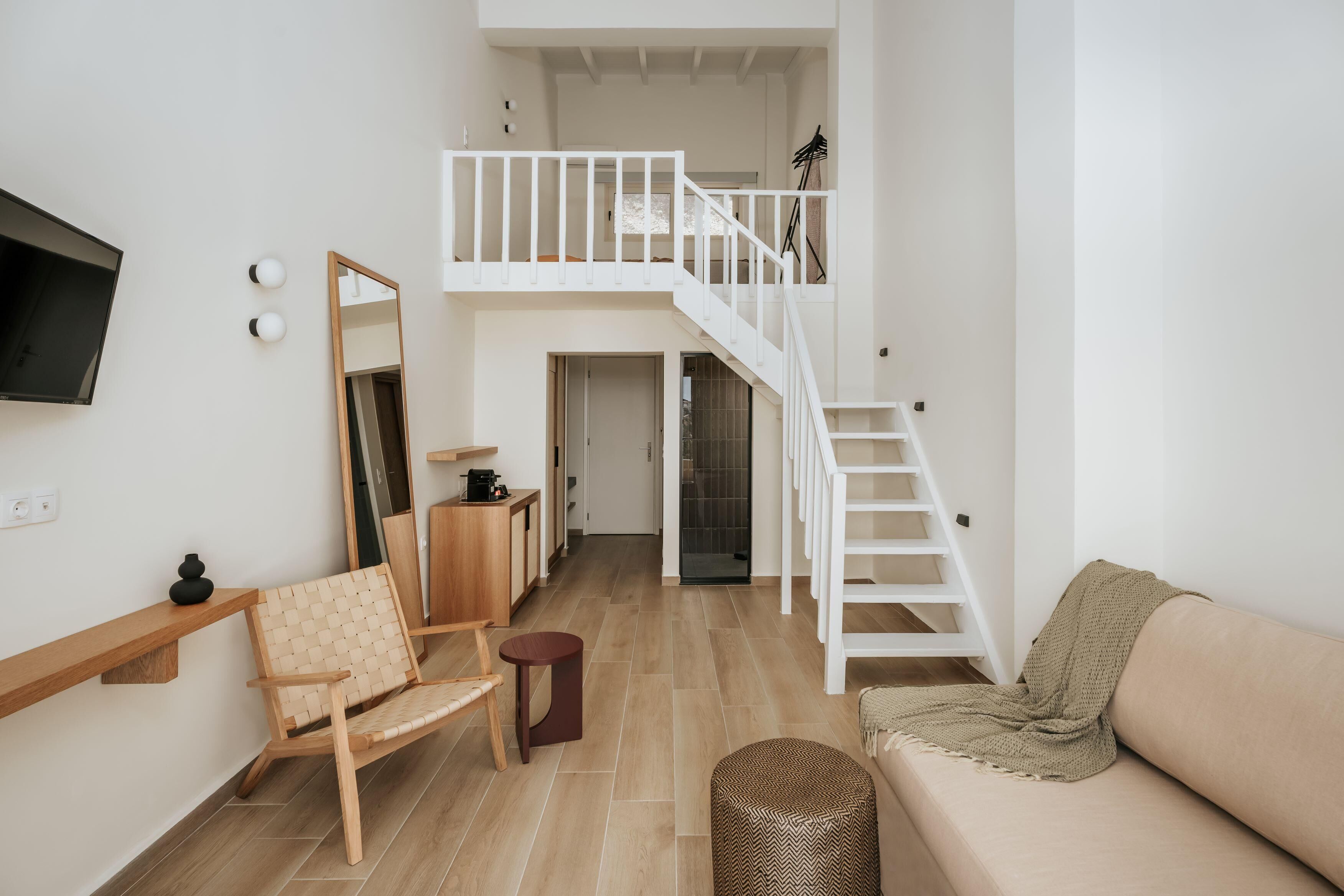 A spacious living area with a high ceiling, a staircase, a sofa, a chair, a small table, and a TV mounted on the wall.
