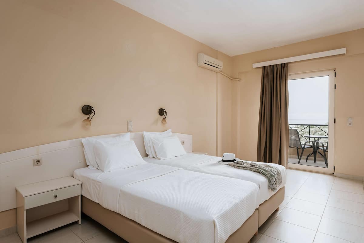 A hotel room with a double bed, a nightstand, an air conditioning unit, and a balcony with a table and chairs.