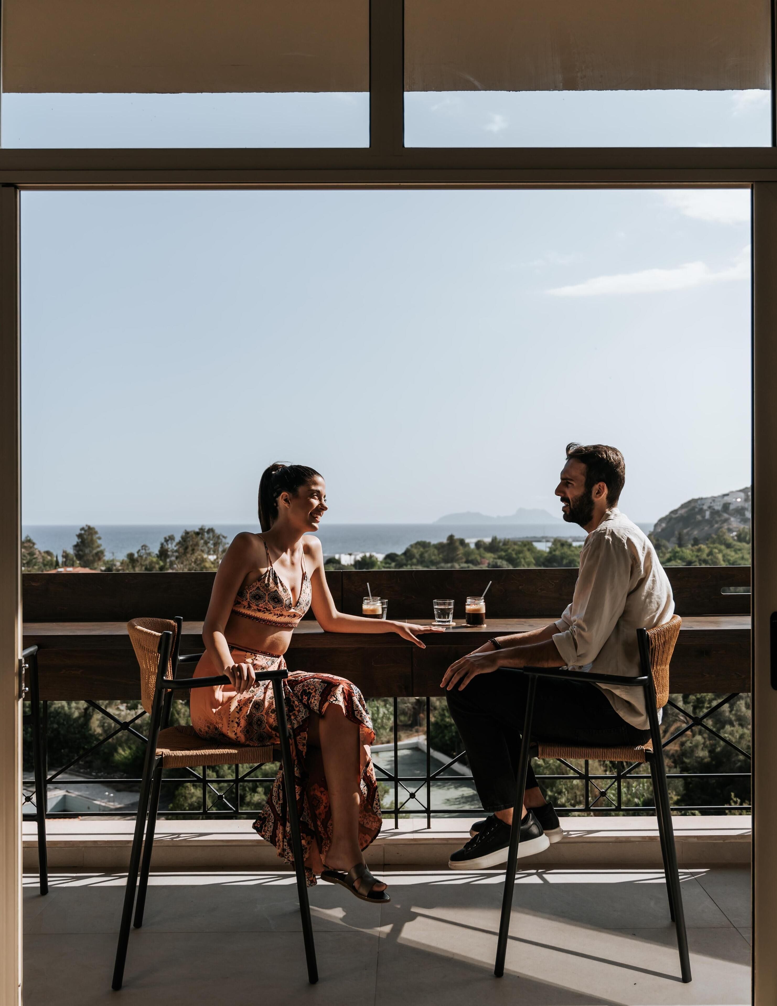 Two people seated at a table with drinks, overlooking a scenic view of mountains and trees.