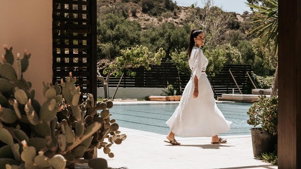 Una persona con vestido blanco caminando hacia un área de piscina con techo de madera y plantas de cactus.