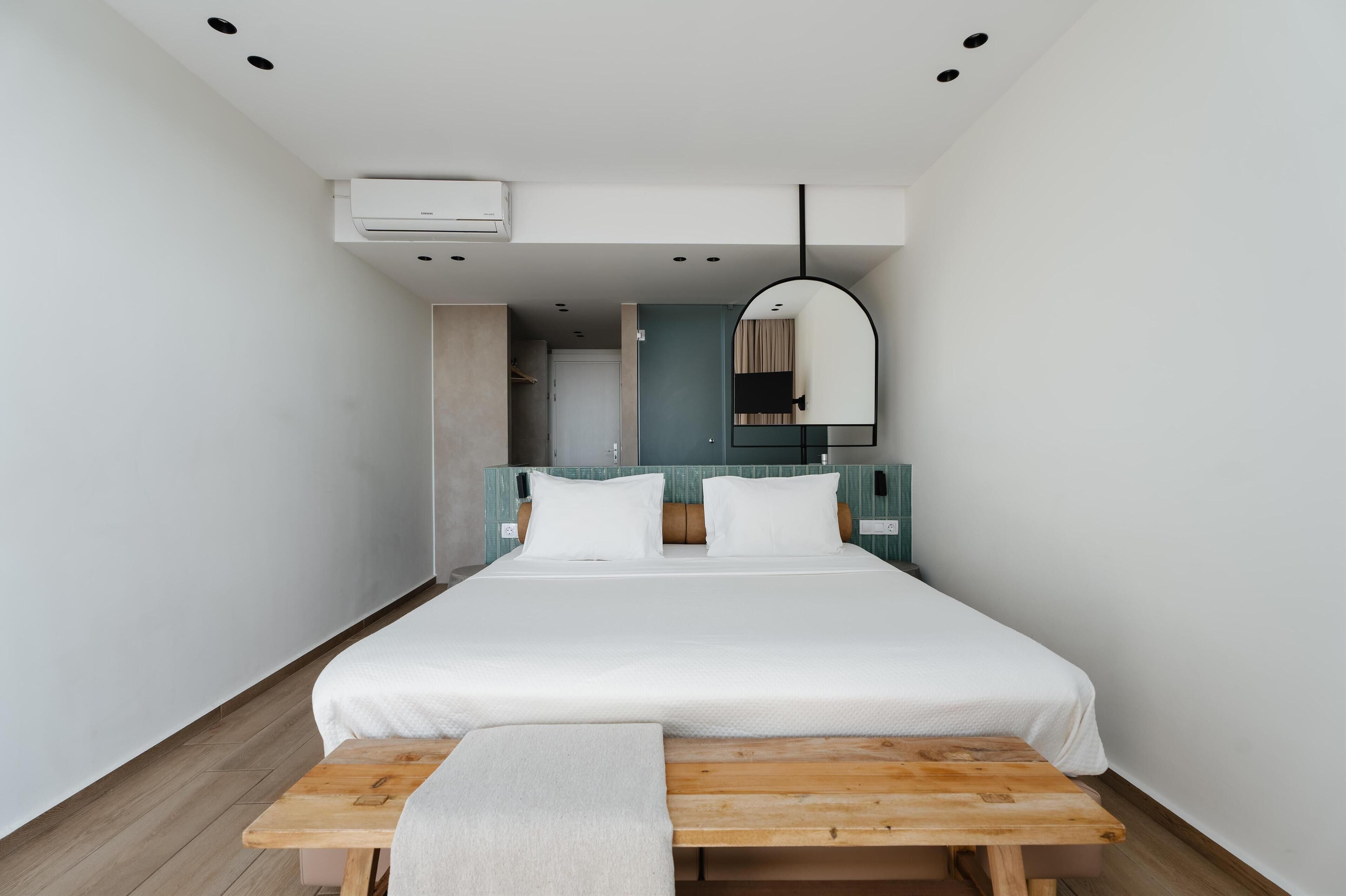 A modern hotel room with a large bed, a wooden bench, a mirror, and an air conditioning unit.
